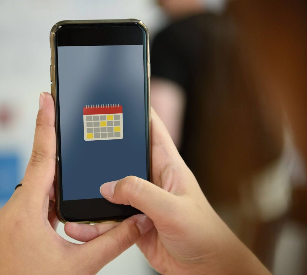 A person holds a smartphone displaying a calendar icon on the screen, with their thumb positioned to tap or swipe, representing the best daily calendar app.