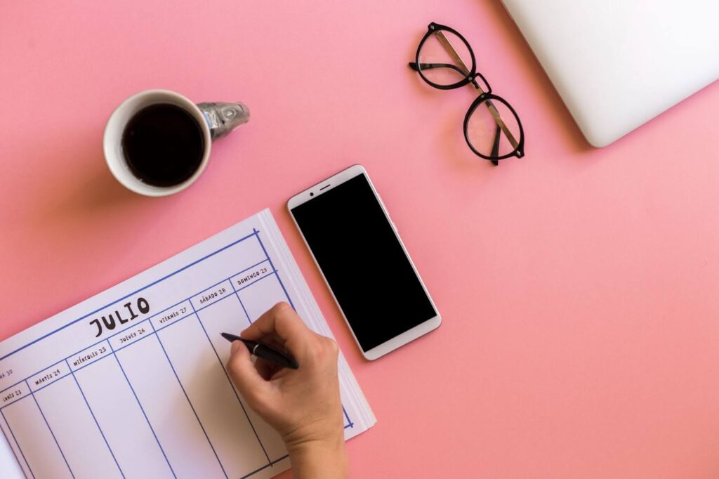 Handwriting in a physical planner for July beside a smartphone, glasses, and coffee, showcasing one of the best daily planners for organized planning.