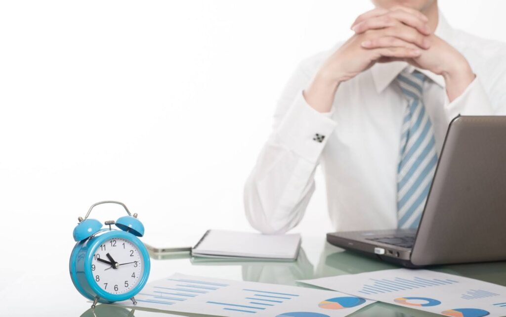 businessman sitting at a desk with a blue alarm clock, charts, and a laptop, symbolizing why time management is important for workplace productivity and performance.